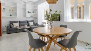 A modern living room with a round wooden dining table, grey upholstered chairs, a grey sofa, white shelves, and a large window with curtains.