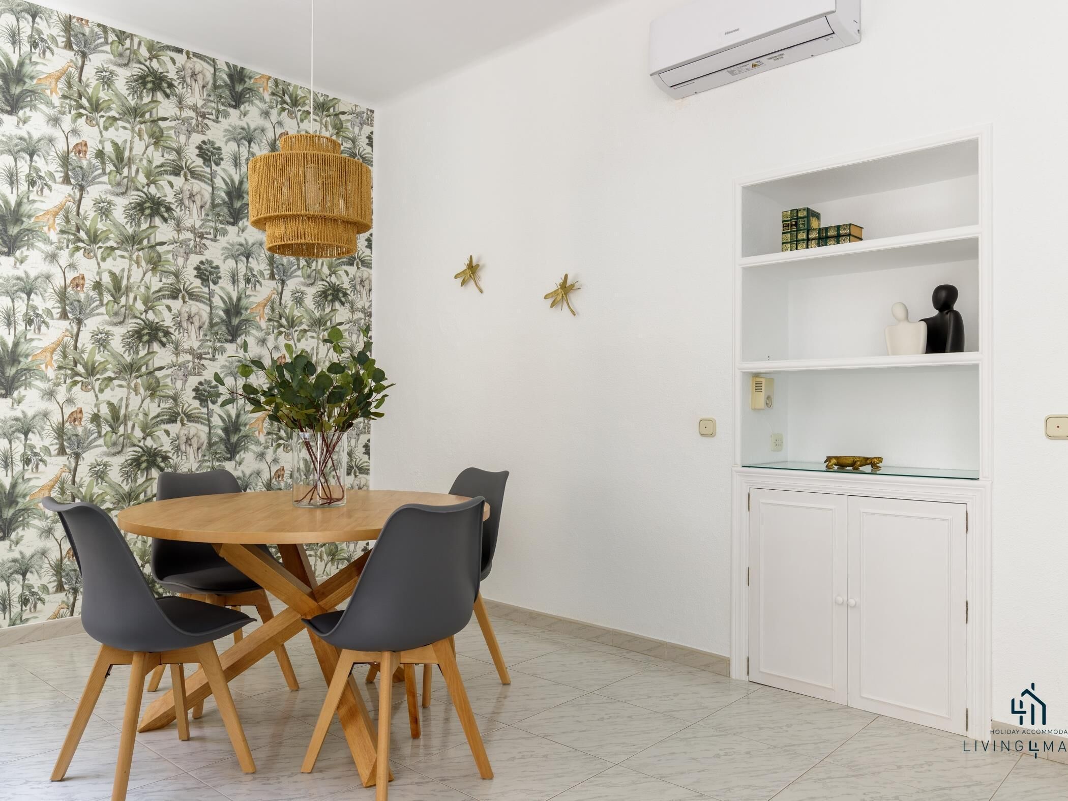A dining area with a round wooden table, black chairs, a woven pendant light, and a wall with a tropical wallpaper.