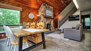Una sala de estar acogedora con techo de madera, chimenea de piedra, una mesa de comedor con sillas y un sofá gris.