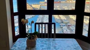 A room with a large window offering a view of a marina with boats and a coastal landscape. - Ertzilla apartament - Sea view. (Bermeo)