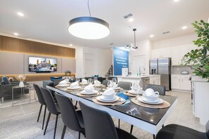 Ett modernt matområde med ett långt bord dukat för en måltid, ett köksområde med vitvaror i rostfritt stål och ett vardagsrum med en platt-TV.