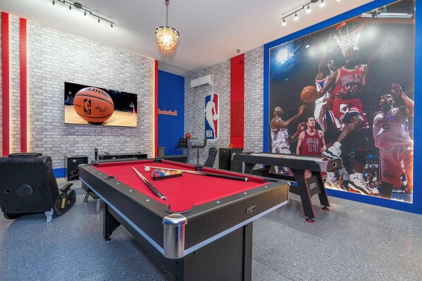Ett biljardbord med en röd filtduk, en tv-skärm på väggen som visar en basketbollmatch och en stor NBA-logotyp på väggen.