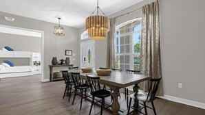 A spacious dining area with a large wooden table and black chairs, a hanging wicker chandelier, and a view of greenery through a large window with curtains.