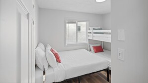 Une chambre avec un lit superposé, un lit simple, des murs blancs, une fenêtre avec des stores et un luminaire au plafond.
