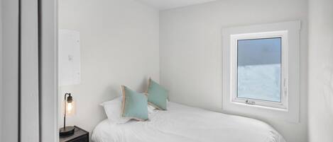 Un dormitorio con cama, almohadas, una mesita de noche con lámpara, una ventana y un interruptor de luz en la pared.