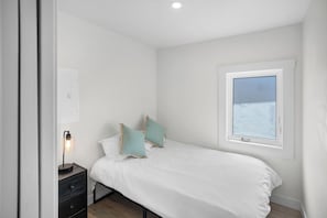 A bedroom with a bed, pillows, a bedside table with a lamp, a window, and a wall-mounted light switch. - Charming Suite with Kitchenette & Shared BBQ, Dock, Firepit (Lake Cowichan)