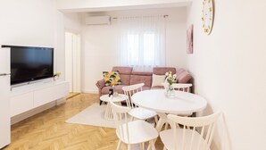 A modern living room with a flat-screen TV, a sofa, a dining table with chairs, and a large window with sheer curtains.