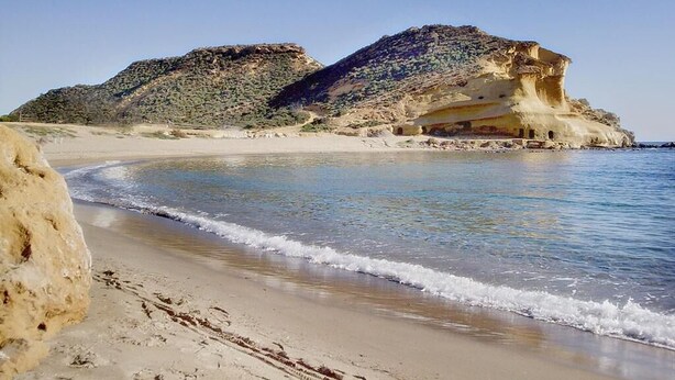 Ein Strand mit klarem Wasser, einem sandigen Ufer und Felsformationen im Hintergrund.