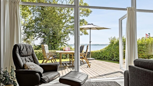 Ein modernes Wohnzimmer mit einem großen Glasfenster, das auf eine Terrassenanlage nach außen führt. Im Innenbereich befindet sich eine Ledersofa, ein Couchtisch aus Holz und ein kleiner Hocker. Draußen eine Holzterrasse mit Tisch und Stühlen sowie einem Sonnenschirm.