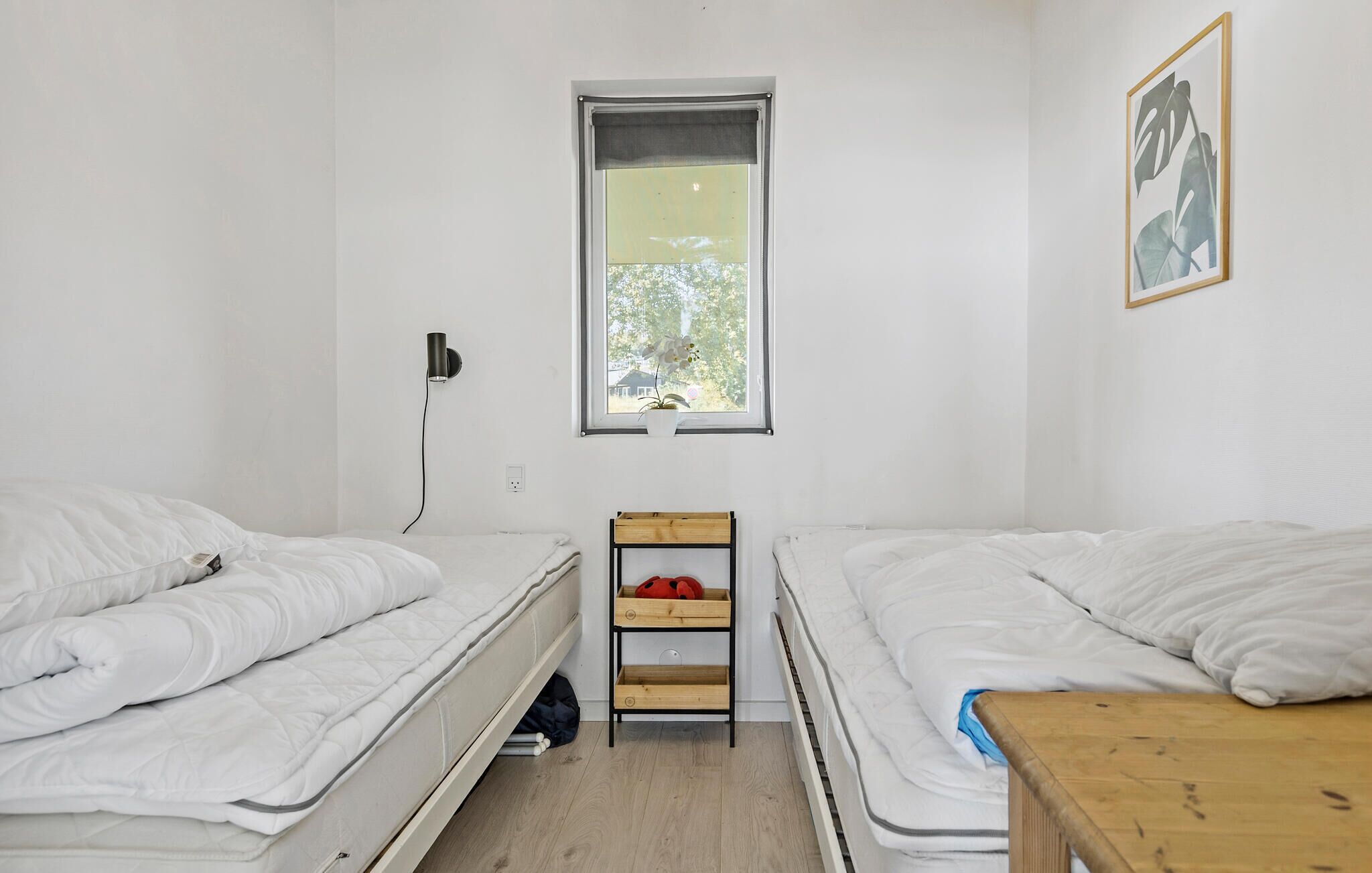 Una habitación pequeña y limpia con dos camas, una ventana con vistas, una lámpara de pared y un estante de madera.
