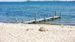 Ein sandiger Strand mit einer hölzernen Pier, die sich in ruhiges, blaues Wasser erstreckt.