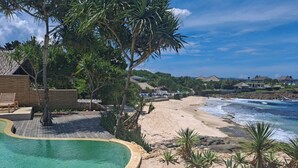 Piscina rodeada por uma praia de areia, com vista para o oceano e casas ao fundo.