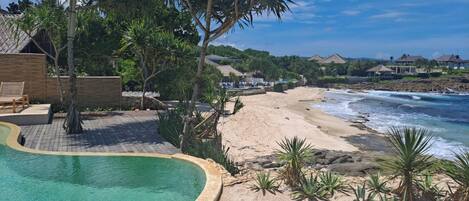A pool surrounded by a sandy beach, with a view of the ocean and houses in the background.