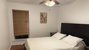 Une chambre à coucher comprenant un lit, une table de chevet, un ventilateur de plafond, une fenêtre avec des stores et une œuvre d’art accrochée au mur.