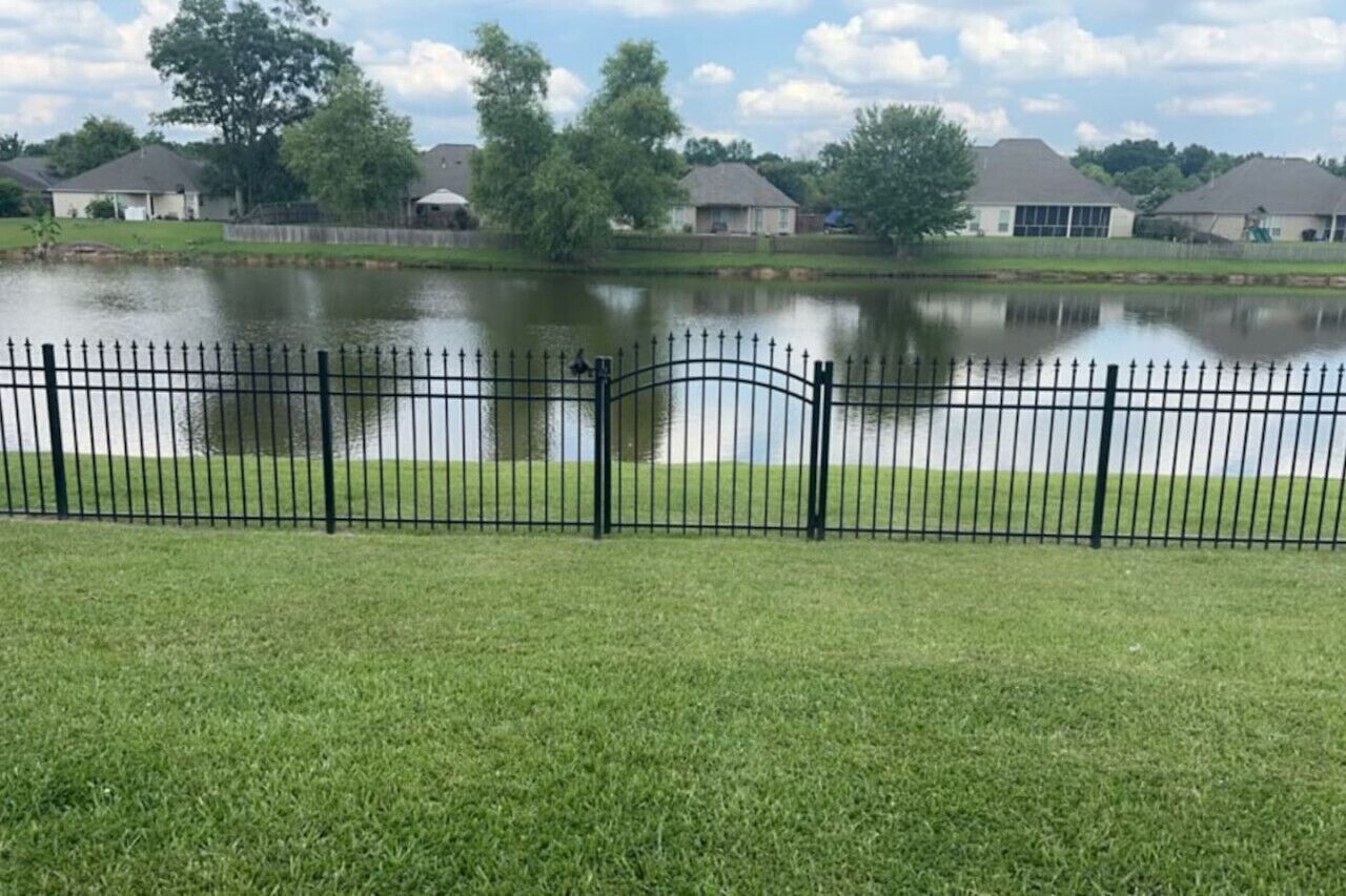 A fenced grassy area with a body of water and houses in the background.