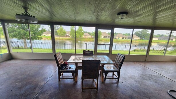 Une terrasse couverte avec une table et des chaises, de grandes fenêtres offrant une vue sur un lac et des maisons.