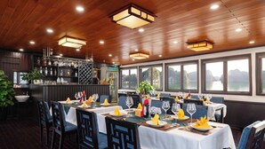 A restaurant with wooden ceiling, tables set with white tablecloths, blue chairs, and yellow napkins. - Garden Bay Legend 2 cruise (Bai Chay)