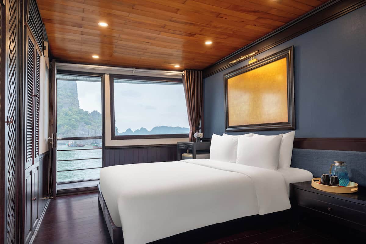 A hotel room with a large bed, a nightstand, a window with a view of the sea, and a wooden ceiling.