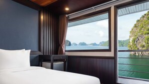 A hotel room with a large bed, a nightstand, a small table, and a window offering a view of a body of water and a mountainous landscape. - Garden Bay Legend 2 cruise (Bai Chay)