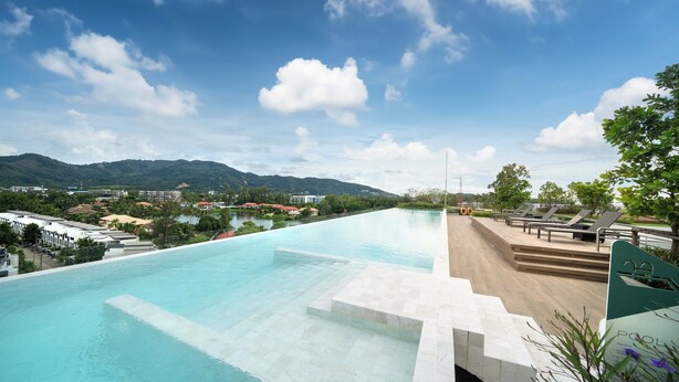 A large outdoor swimming pool with clear blue water, surrounded by steps and lounge chairs, with a view of mountains and buildings in the background. - Laguna Skypark B2512 (Choeng Thale)
