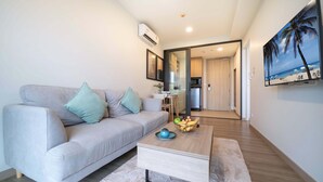 A modern living room with a grey sofa, a coffee table with fruits, and a kitchen in the background. - Laguna Skypark B2512 (Choeng Thale)