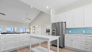 Une cuisine moderne avec des meubles blancs, un réfrigérateur en acier inoxydable et une vue sur la plage grâce à de grandes fenêtres.