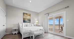 A bedroom with a bed, a nightstand, a lamp, a wicker chair, a painting, and a balcony with a view.
