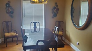Une salle à manger avec une table en bois, quatre chaises, un lustre et un grand miroir au mur.