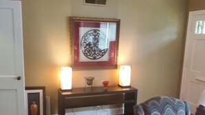 Un salon doté d’un plancher en bois, d’une œuvre d’art encadrée au mur, d’une petite table avec deux bougies et d’un fauteuil recouvert d’un tissu à motifs.