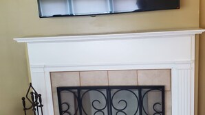 Un salon doté d’un téléviseur à écran plat fixé au-dessus d’une cheminée blanche, d’un paravent décoratif en métal et d’un ventilateur de plafond.