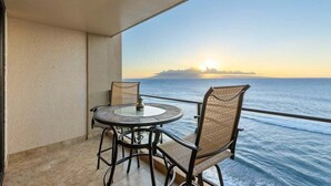 A balcony with a round table and two chairs, overlooking a body of water with a sunset in the background. - Perch - Oceanfront Tranquility: 1BR Condo w/ Views (Lahaina)