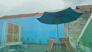 An outdoor patio with a table and chairs, a large umbrella, and a view of a blue wall and tiled roof.