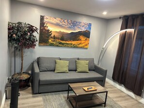 A living room with a grey sofa, two green cushions, a coffee table with a remote control, a potted plant, and a painting of a mountain landscape.
