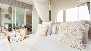 A modern bedroom with a large bed, white bedding, and a view of a cityscape through large windows. - Family Suite Sea view 2Bedroom  (Pattaya)