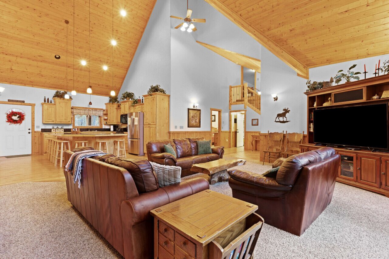 A spacious living area with a high ceiling, wooden beams, a large TV, and a kitchen in the background.