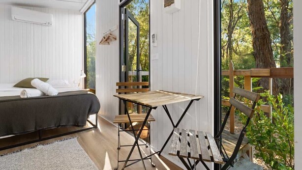A room with a bed, a small table, and a bench. There is a window with a view of trees. - Orange 2 by Tiny Away (Woori Yallock)