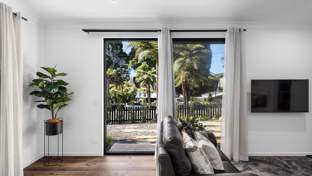 A modern living room with a leather sofa, a potted plant, and a sliding glass door leading to a backyard with trees and a fence.