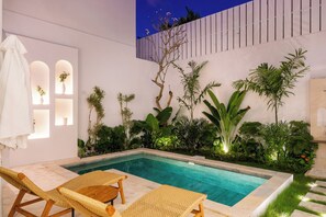Uma área junto à piscina com uma piscina retangular, espreguiçadeiras de vime e diversas plantas tropicais.
