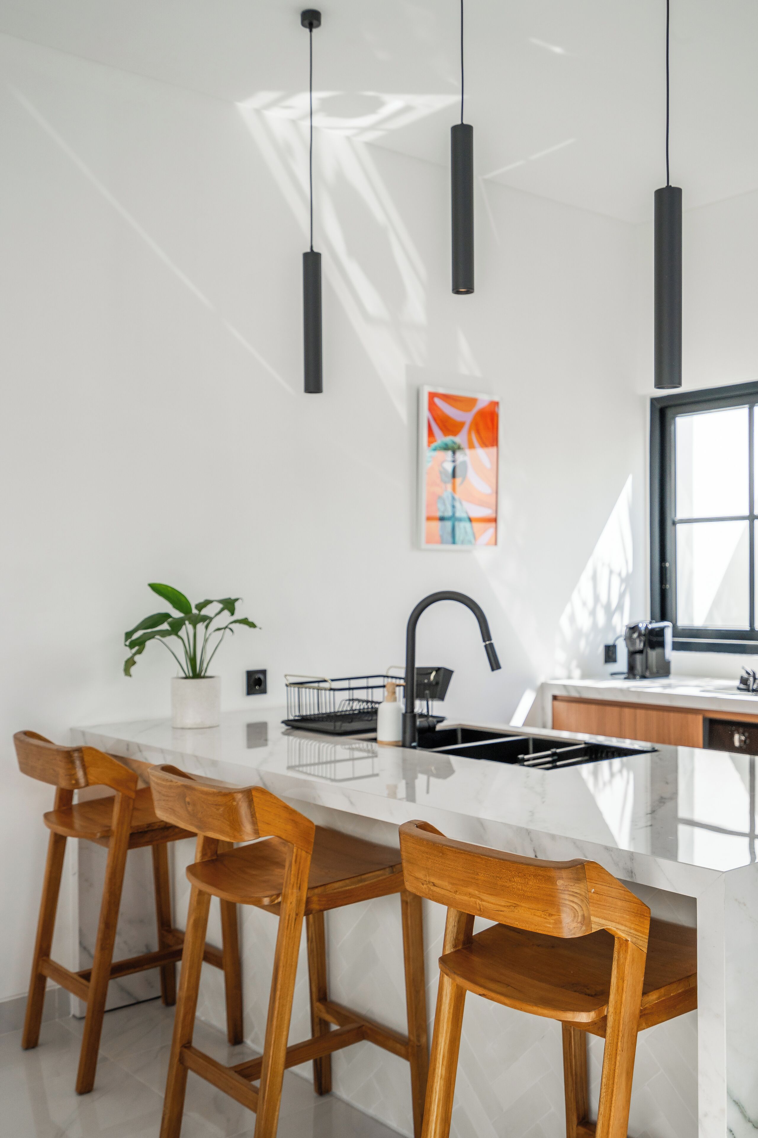 A modern kitchen with a marble countertop, wooden bar stools, pendant lights, a potted plant, and a window.