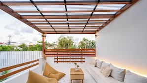 A covered outdoor seating area with a sofa and cushions, a small table with a vase, and a view of greenery and buildings in the background. - Bucu Villa by Betterplace (Kerobokan)