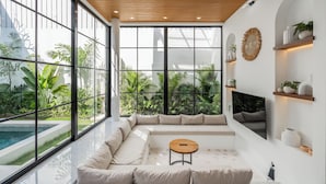 A modern living room with large windows, a sectional sofa, a TV, and a small table. - Bucu Villa by Betterplace (Kerobokan)