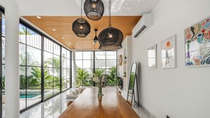 A spacious dining area with a wooden table and chairs, large windows offering a view of a swimming pool and greenery, and modern pendant lighting. - Bucu Villa by Betterplace (Kerobokan)