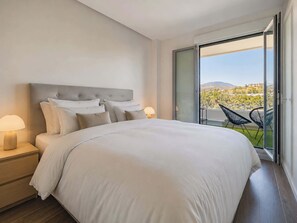Une chambre d’hôtel moderne dotée d’un grand lit, de deux tables de chevet avec des lampes, d’une porte-fenêtre coulissante donnant sur un balcon avec des chaises, et offrant une vue sur la ville.