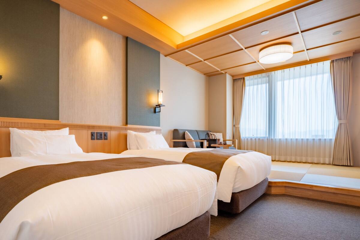 A hotel room with two beds, a nightstand, a lamp, and a window with curtains.