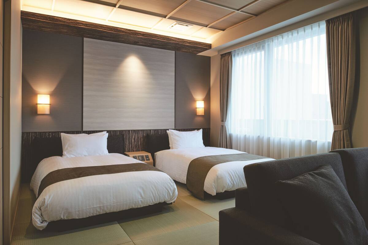 A hotel room with two beds, a sofa, a wall-mounted TV, and a window with curtains.