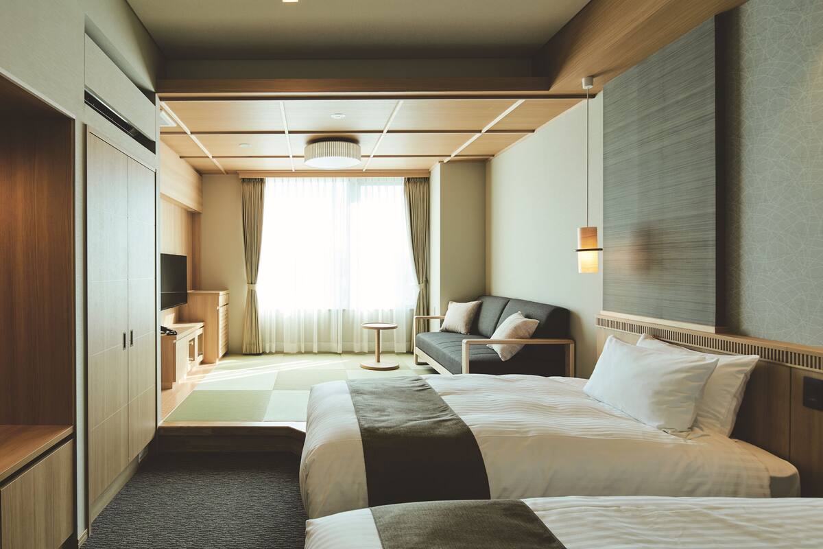 A hotel room with two beds, a sofa, a TV, and a large window with curtains.