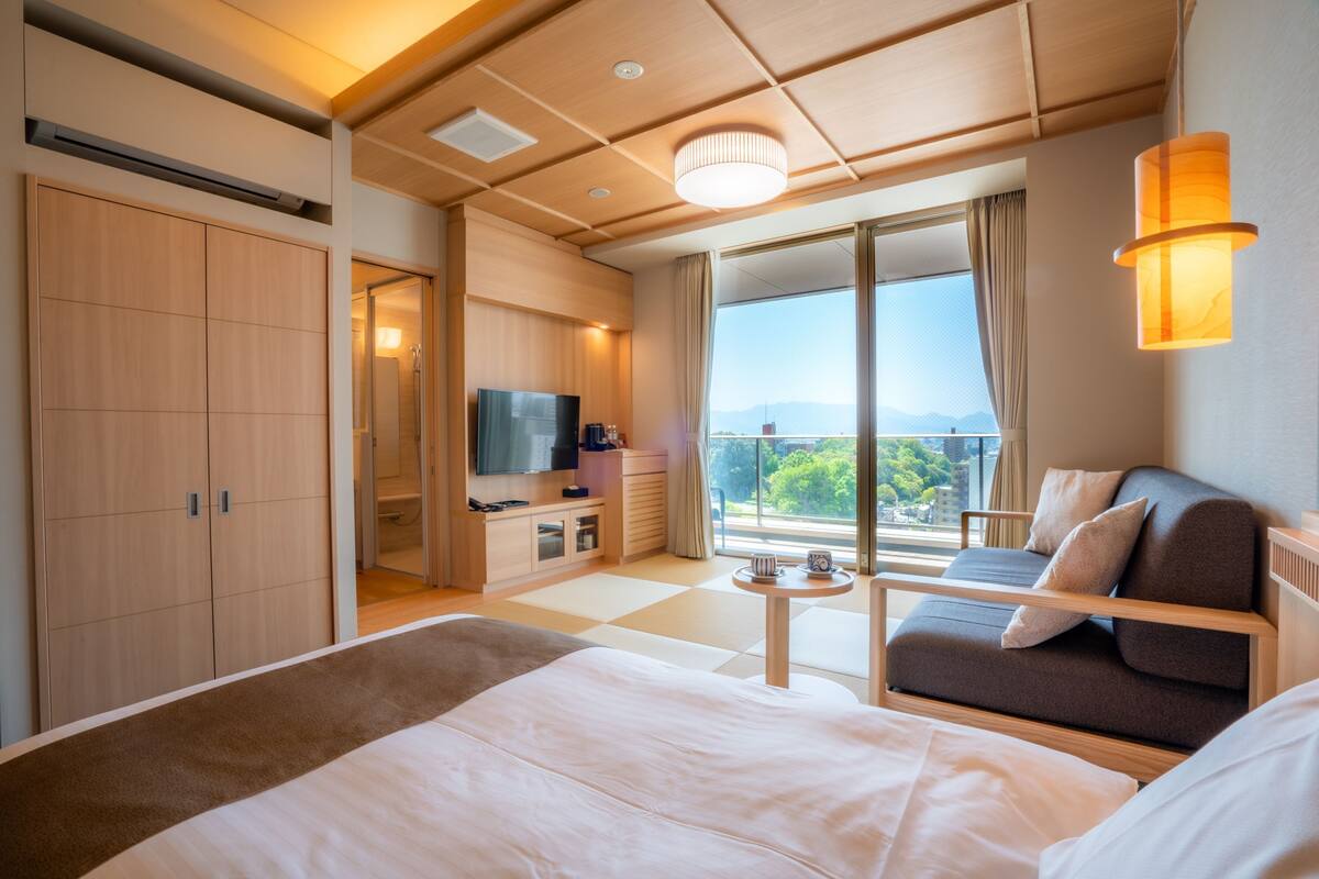 A modern hotel room with a large bed, a sofa, a TV, a small table, and a balcony with a view.