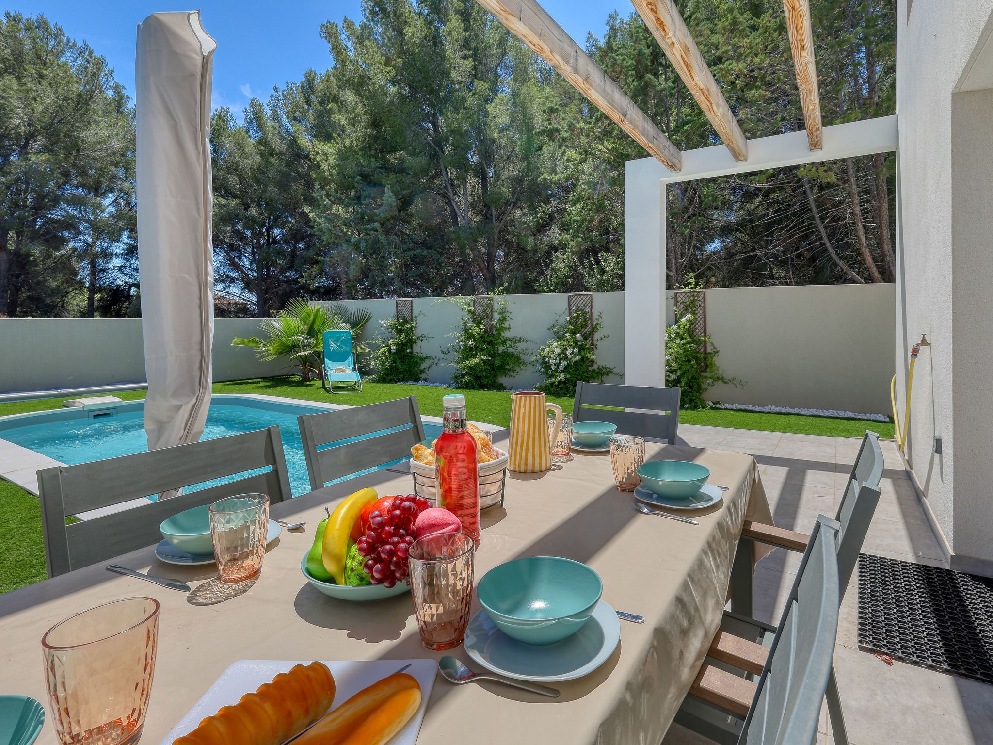 An outdoor dining area with a pool, chairs, and a table set with fruits, glasses, and a bottle.