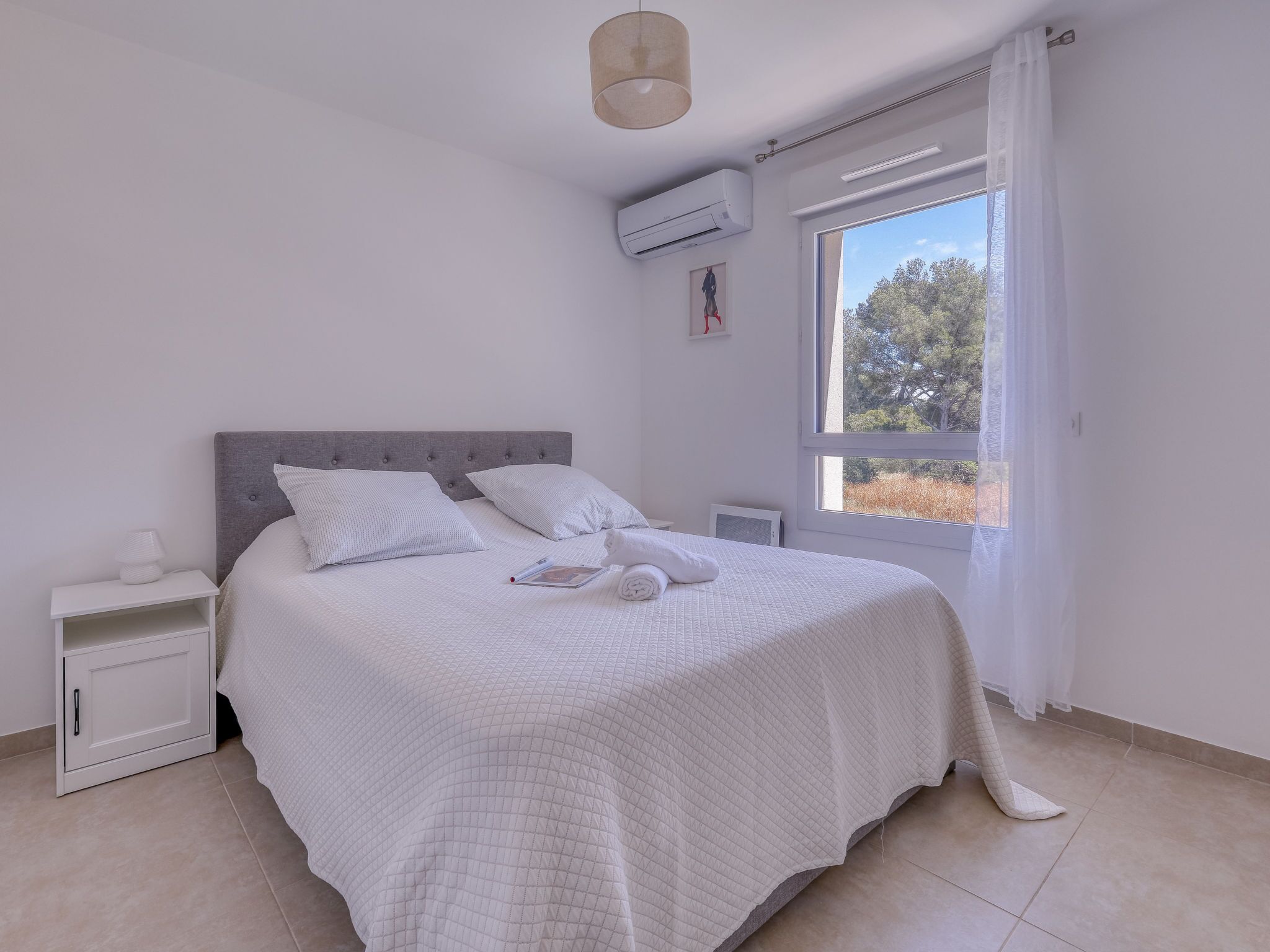 A bedroom with a bed, a nightstand, a window with a view of trees, and an air conditioning unit.