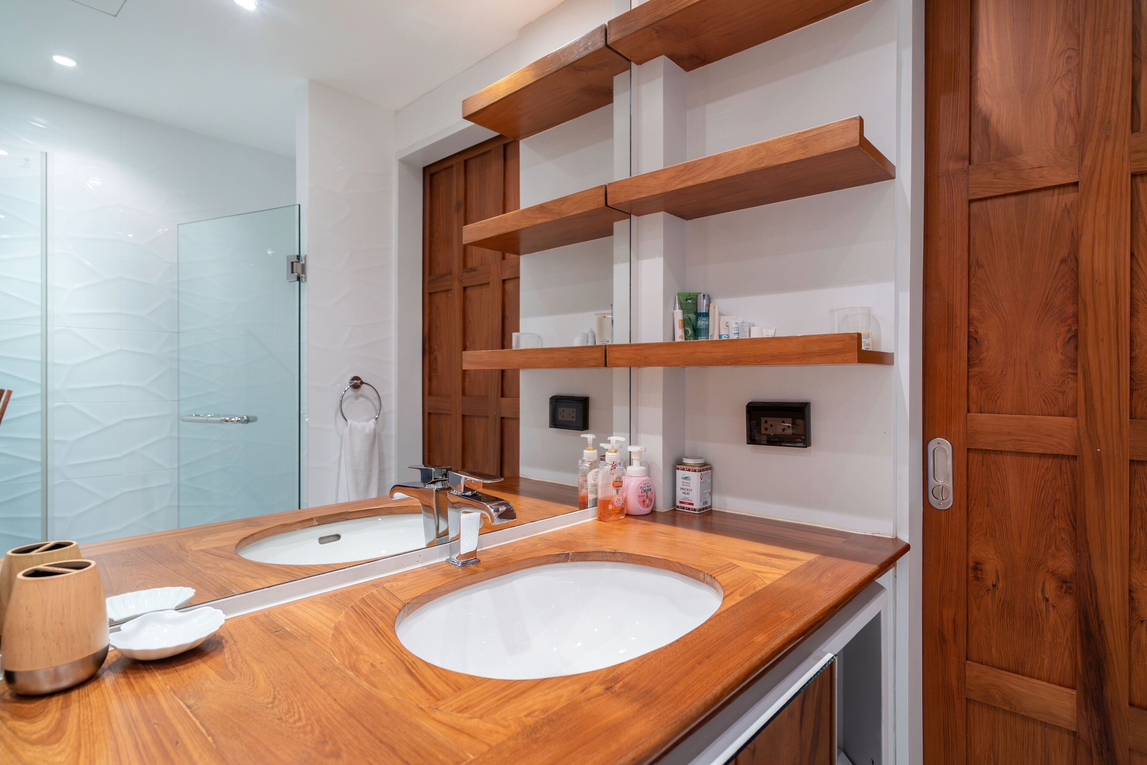 A modern bathroom with a wooden vanity, two sinks, and built-in shelves above the mirror.